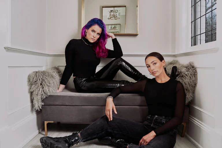 One woman with long purple hair sitting on a couch and one woman with tied back long brown hair and black combat boots sitting on the ground.