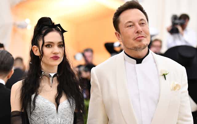 Elon Musk in a white dress suit accompanied by his and his partner Grimes in an embroidered dress.