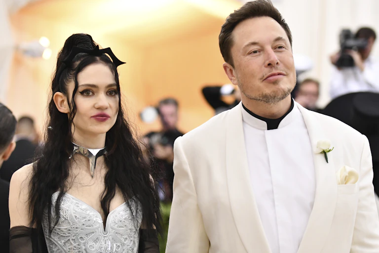 Elon Musk in a white dress suit accompanied by his and his partner Grimes in an embroidered dress.