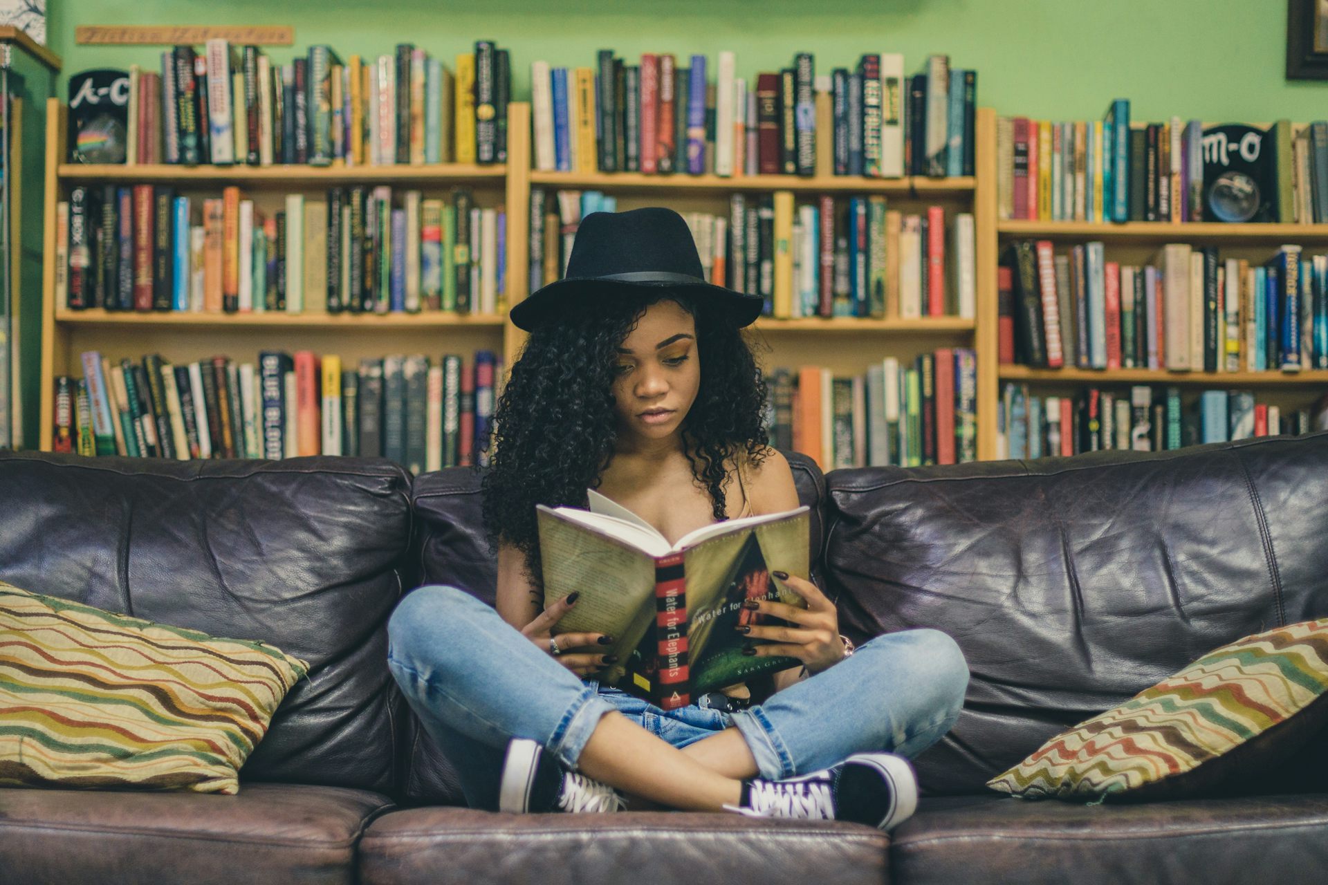 A woman in a hat sitting cross legged on a couch reading.