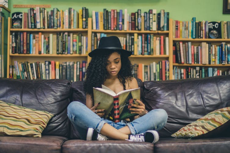 A woman in a hat sitting cross legged on a couch reading.