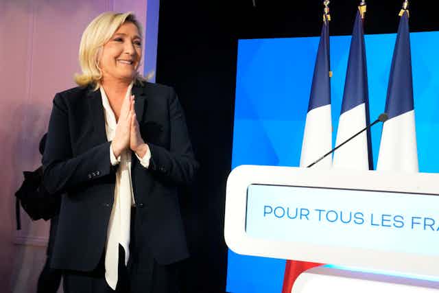 Marine Le Pen, les mains jointes, à côté de drapeaux français.