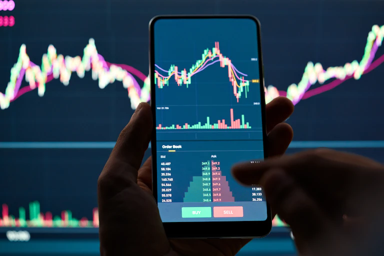 a person's hands holding a mobile phone in front odf a screen, both showing stocks