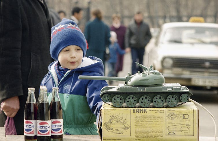A white boy with a blue stocking hat and coat looks at a toy tank on a box on a table next to several glass Pepsi bottles