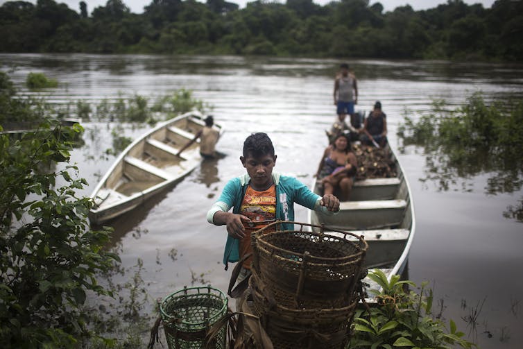 A young person drops off baskets while people wait behind him in a narrow boat holding manioc, an edible root.