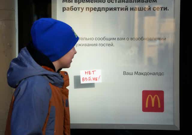 A white boy in a blue hat and coat walks past sign with a McDonalds golden arch logo, an explanation in Russia of why the story is closed and a small note posted to the glass reading, in russia, no war!