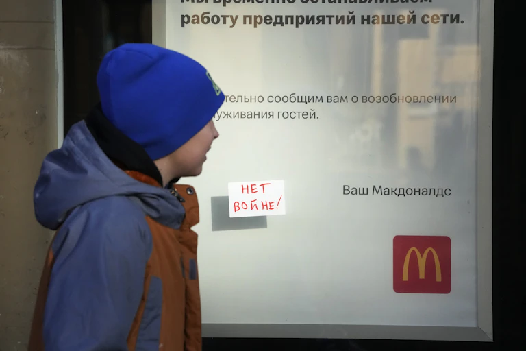 A white boy in a blue hat and coat walks past sign with a McDonalds golden arch logo, an explanation in Russia of why the story is closed and a small note posted to the glass reading, in russia, no war!