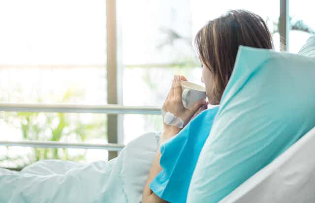 Woman in surgical gown lays in hospital bed sipping tea.