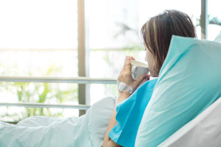 Woman in surgical gown lays in hospital bed sipping tea.