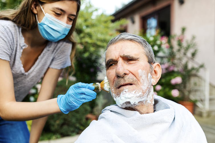 Woman in mask shaves elderly man.