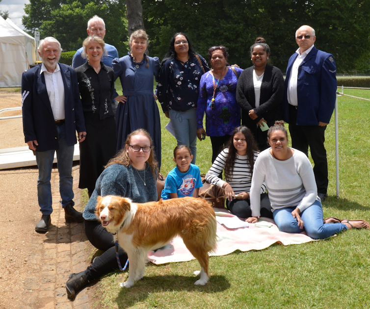 A group of 12 people in period costume with a dog posing on a green lawn.