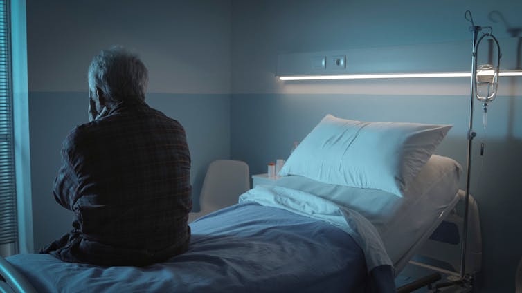 Man sits on the edge of a hospital bed in the dark.