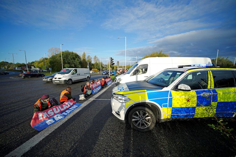 Insulate Britain protesters seated on a motorway and holding banners, with two police vans parked in front of them