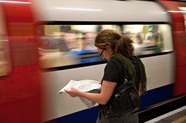 A woman reads a newspaper as a blurred Tube train passes at speed