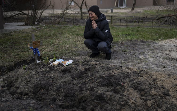 A woman in a black hat and jacket kneels next to a grave.