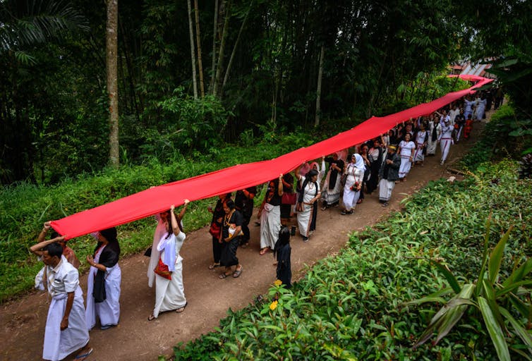 People walk in a long line under a huge red banner along a wooded path.
