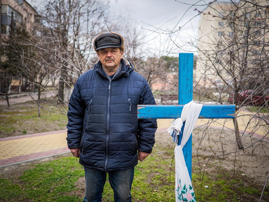 A man in a winter coat and hat stands next to a blue cross over a grave.