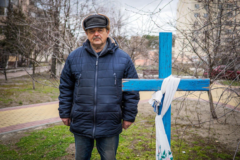 A man in a winter coat and hat stands next to a blue cross over a grave.