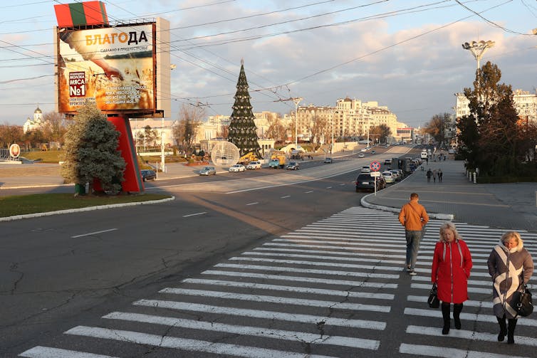 Emberek sétálnak át az átkelőn egy szinte üres belvárosban, egy nagy karácsonyfával és épületekkel a háttérben