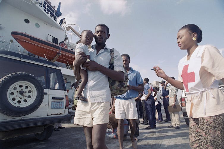Haitian men walking in a line wearing T-shirts and shorts, next to a ship, while a woman looks on.