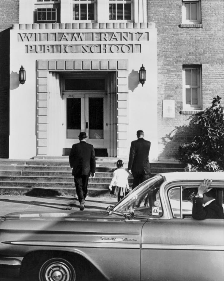 Two men dressed in dark suits are seen walking up stairs to a school with a little black girl.