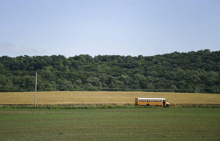 A School Bus Drives Along A Road Past A Green Field, With Woods In The Background
