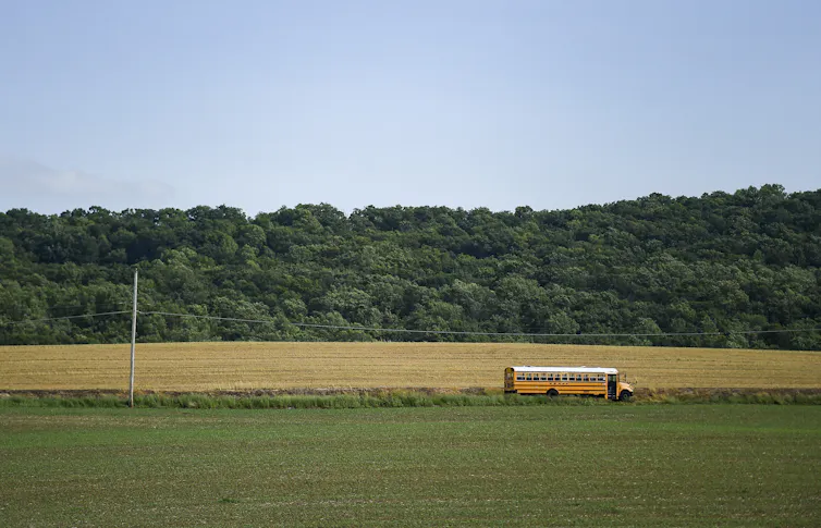 A school bus drives along a road past a green field, with woods in the background