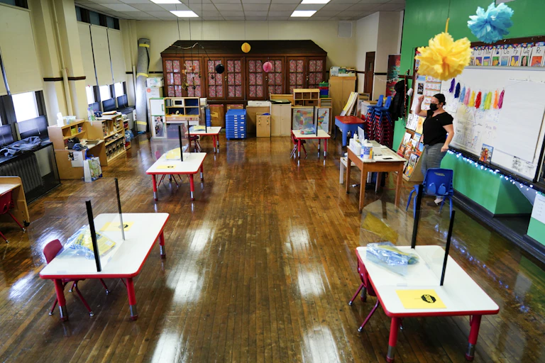 A teacher stands in front of an empty classroom, speaking to a computer screen.