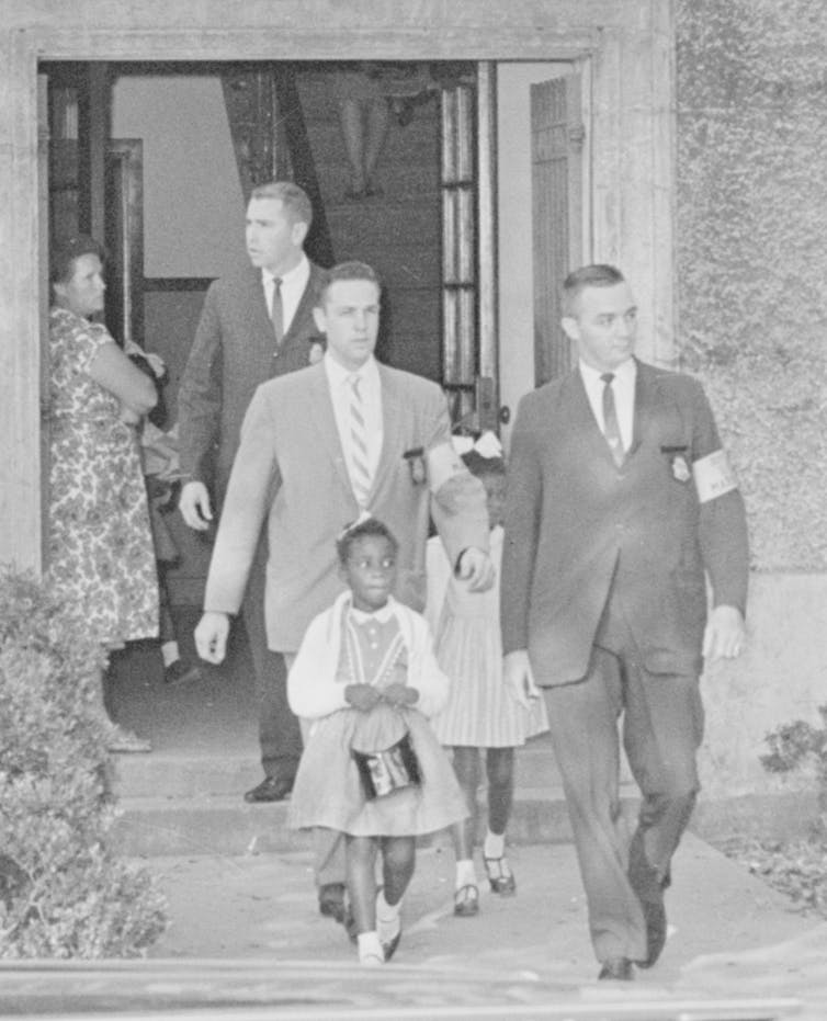 Three white men dressed in business suits and wearing arm bands are escorting three black girls from school.