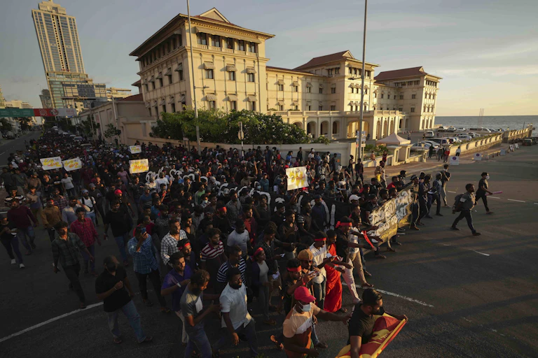 A large crowd, some holding signs, marches past a building on the seashore.