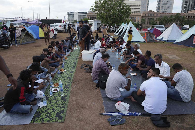 People sit on long blankets outside for a meal.