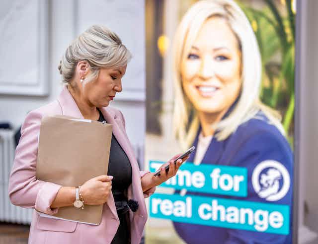 Sinn Fein's Deputy Leader Michelle O'Neill checking her phone in front of an election campaign poster bearing her face.