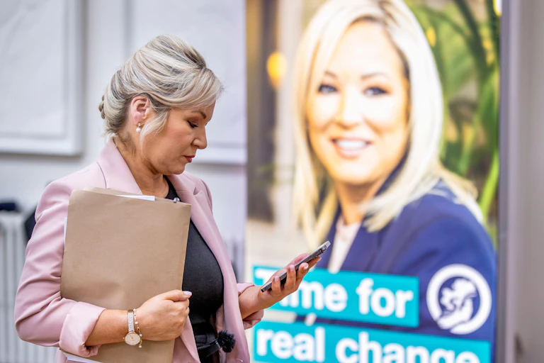 Sinn Fein's Deputy Leader Michelle O'Neill checking her phone in front of an election campaign poster bearing her face.