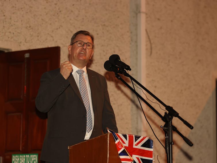 DUP leader Jeffrey Donaldson speaking at a podium bearing the union flag during a rally at night in Northern Ireland.