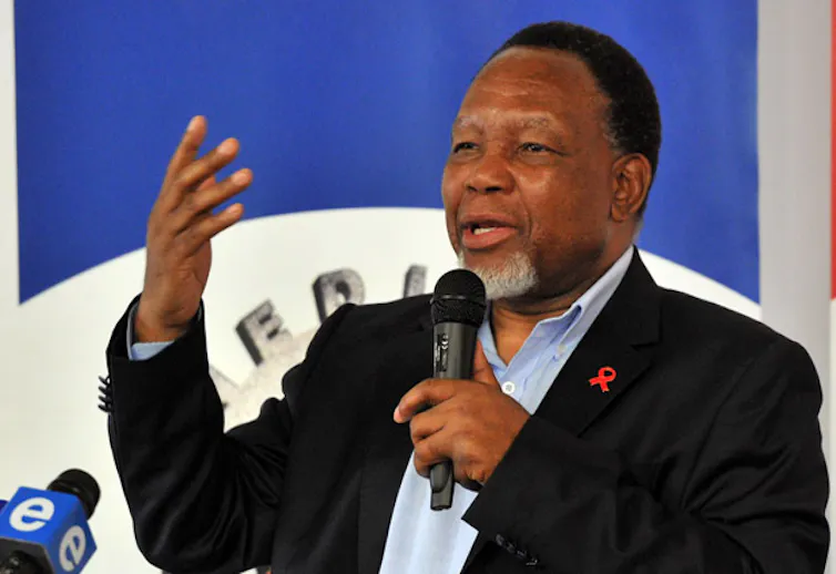 A man makes an open palm gesture with his right had as he speaks into a microphone in his left hand.