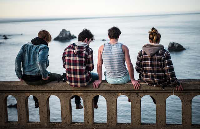 A group of four young people, sitting on a low wall and seen from behind, look out over the ocean.