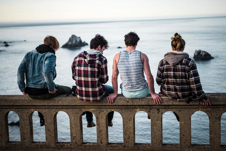A group of four young people, sitting on a low wall and seen from behind, look out over the ocean.