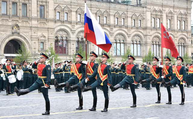 Soldiers marching carrying red, blue and white flag, with a white building behind them.