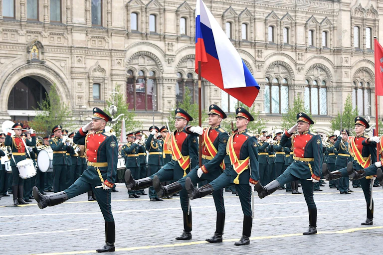 Soldiers marching carrying red, blue and white flag, with a white building behind them.
