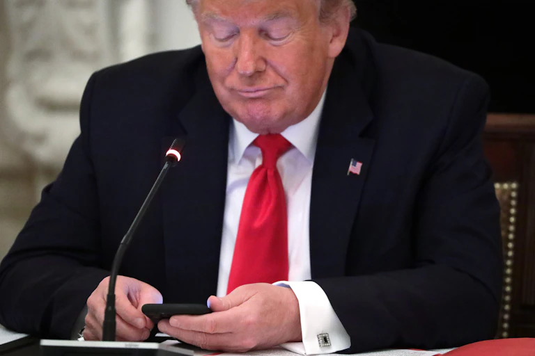 Donald Trump wearing a dark suit and red tie looks at his phone.