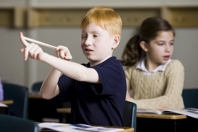 A young boy sitting at a school desk aims a rubber band