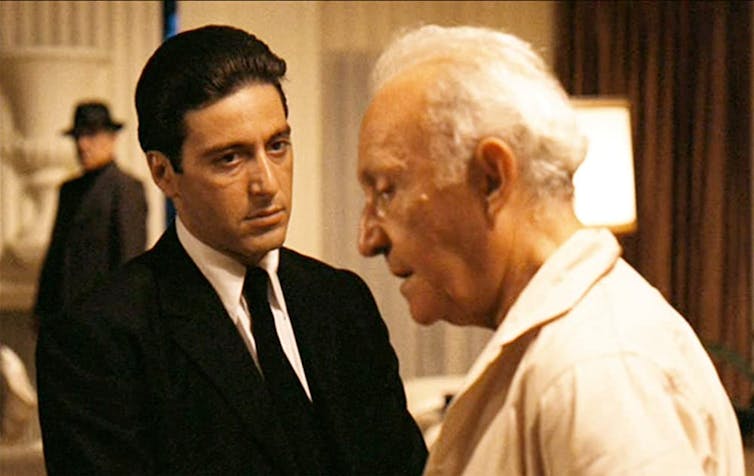 A young man in a suit and tie talks to an older, white-haired man.