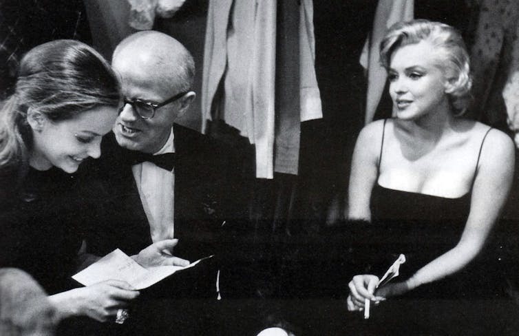 Older man and daughter laughing over a piece of paper while glamorous blonde sits to their side.