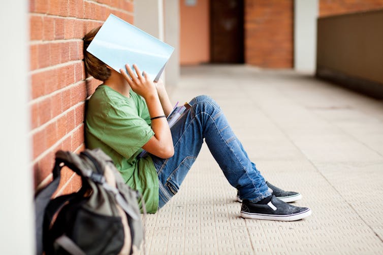 A teenage boy in a green t-shirt and jeans sits on the ground holding a book over his head.