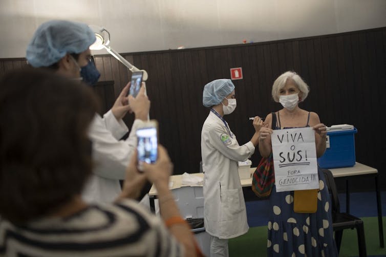 A Brazilian woman receives a vaccination jab while holding a sign denouncing the government’s inaction