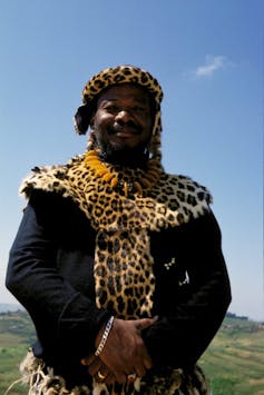 A man wearing a headband made of leopard skin folds his hands on his stomach.