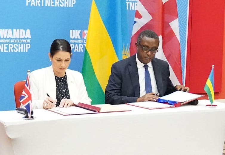 Home Secretary Priti Patel and Rwandan minister for foreign affairs and international co-operation, Vincent Biruta, signing documents at a table in front of their national flags.