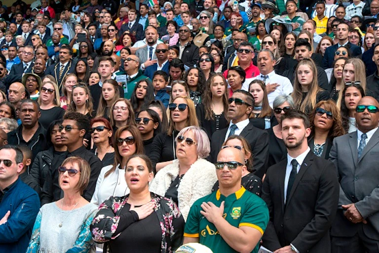 A crowd of men and women, some with their hands to their chests, sings the South African National Anthem.