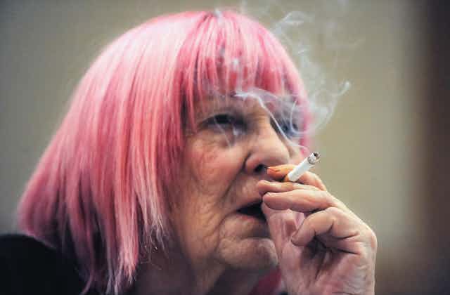 Elderly woman with pink hair smokes a cigarette.