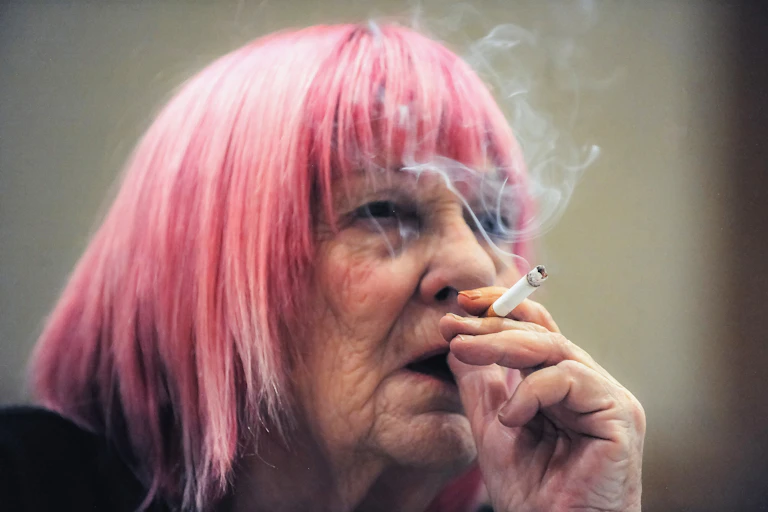 Elderly woman with pink hair smokes a cigarette.